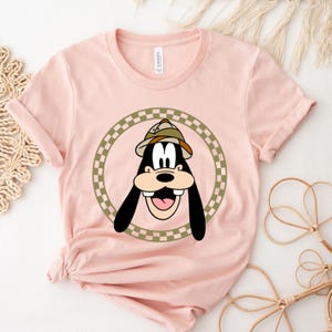 May include: A pink t-shirt with a cartoon image of Goofy wearing a safari hat. Goofy is surrounded by a green and white checkered circle.
