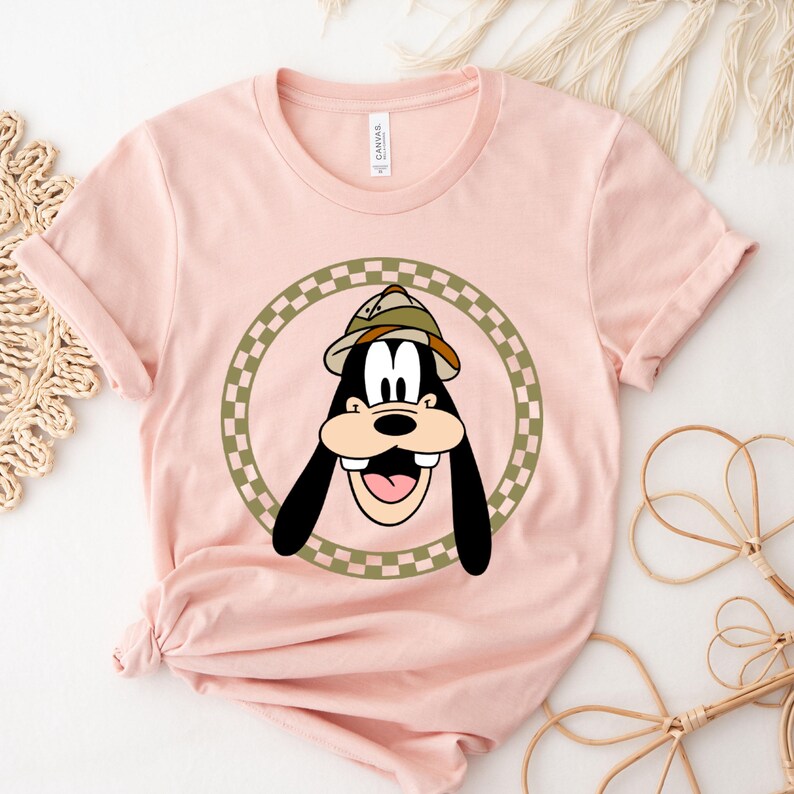 May include: A pink t-shirt with a cartoon image of Goofy wearing a safari hat. Goofy is surrounded by a green and white checkered circle.