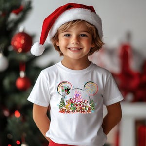 May include: A child wearing a white t-shirt with a colorful Disney Christmas design and a red Santa hat. The shirt features Mickey Mouse ears, a castle, and the word "CHRISTMAS". The background includes a Christmas tree and ornaments.
