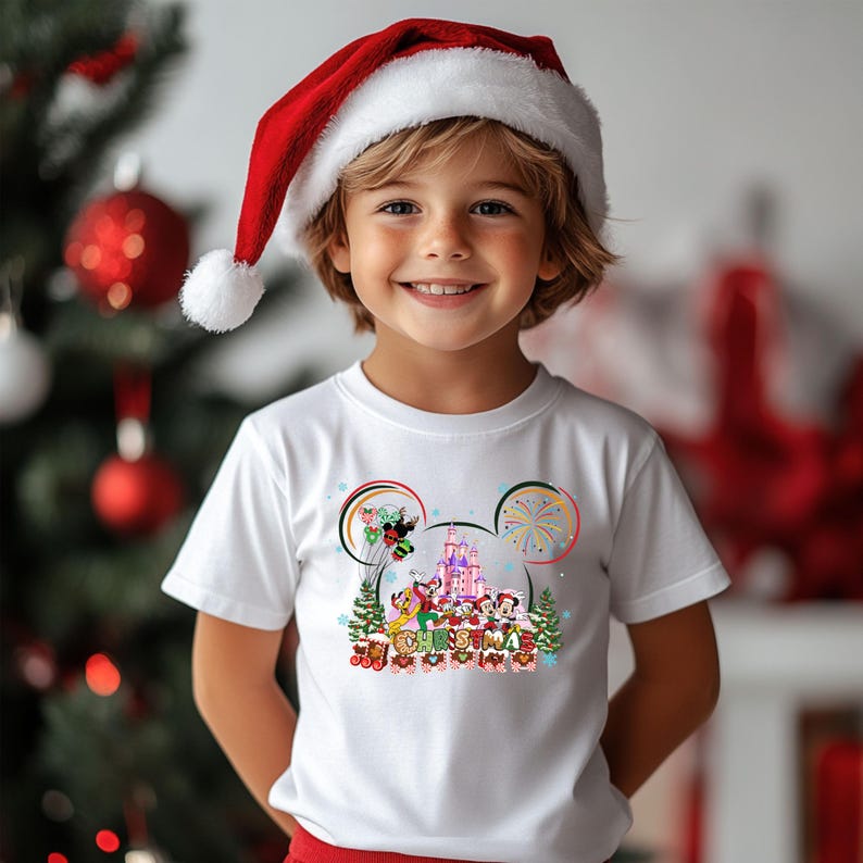 May include: A child wearing a white t-shirt with a colorful Disney Christmas design and a red Santa hat. The shirt features Mickey Mouse ears, a castle, and the word "CHRISTMAS". The background includes a Christmas tree and ornaments.