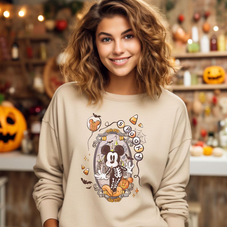May include: Beige sweatshirt featuring a Halloween-themed Mickey Mouse design. The graphic includes Mickey in a skeleton costume, surrounded by candy corn, bats, and the words "Mickey's House". The sweatshirt has long sleeves and a crew neck.