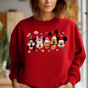 Op de afbeelding: Rood sweatshirt met een afbeelding van Mickey Mouse, Minnie Mouse, Donald Duck, Goofy en Pluto die hartvormige zonnebrillen dragen. De afbeelding bevat ook harten, een sleutel en het woord "Love!"