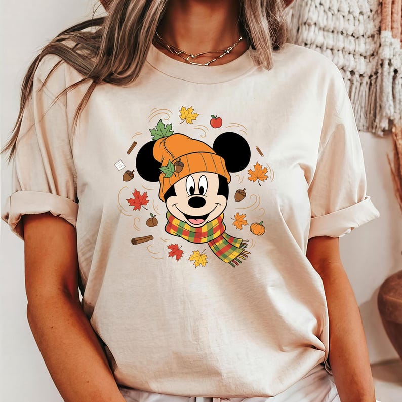 Puede incluir: Camiseta beige con un dise&ntilde;o de Mickey Mouse con un atuendo oto&ntilde;al. Mickey lleva un gorro naranja y una bufanda colorida, rodeado de hojas de oto&ntilde;o, bellotas y una peque&ntilde;a calabaza.