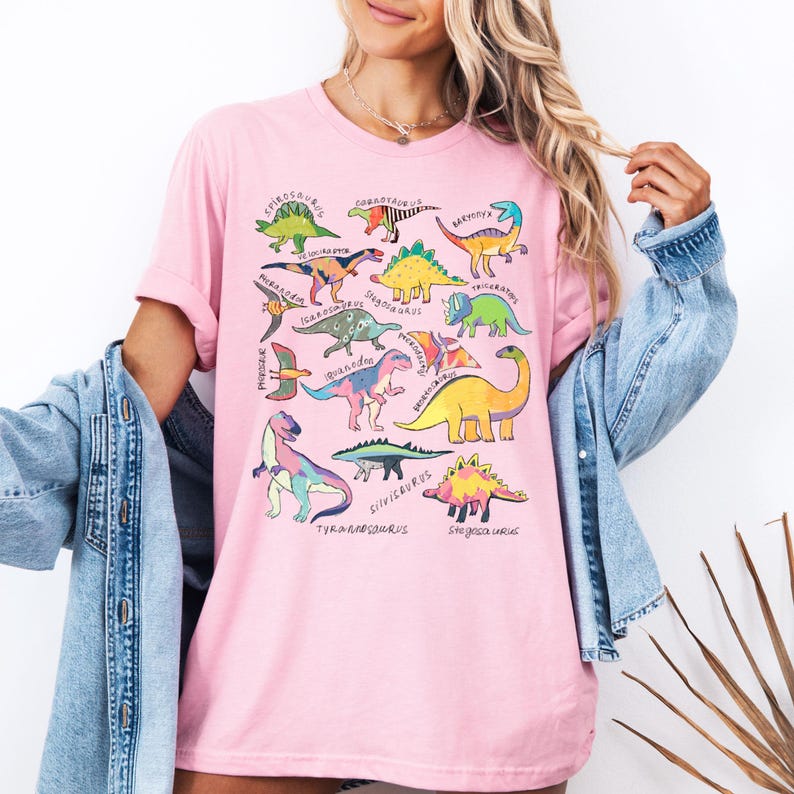 May include: Pink t-shirt with a colorful dinosaur graphic print. The print features various dinosaurs with their names written underneath. The names include:  Stegosaurus, Corythosaurus, Baryonyx, Velociraptor, Miragaia, Iguanodon, Stegoceras, Triceratops, Psittacosaurus,  Guanlong,  Protoceratops,  Brachiosaurus,  Silvicercus,  Tyrannosaurus,  Shunosaurus,  and  Sinosauropteryx.