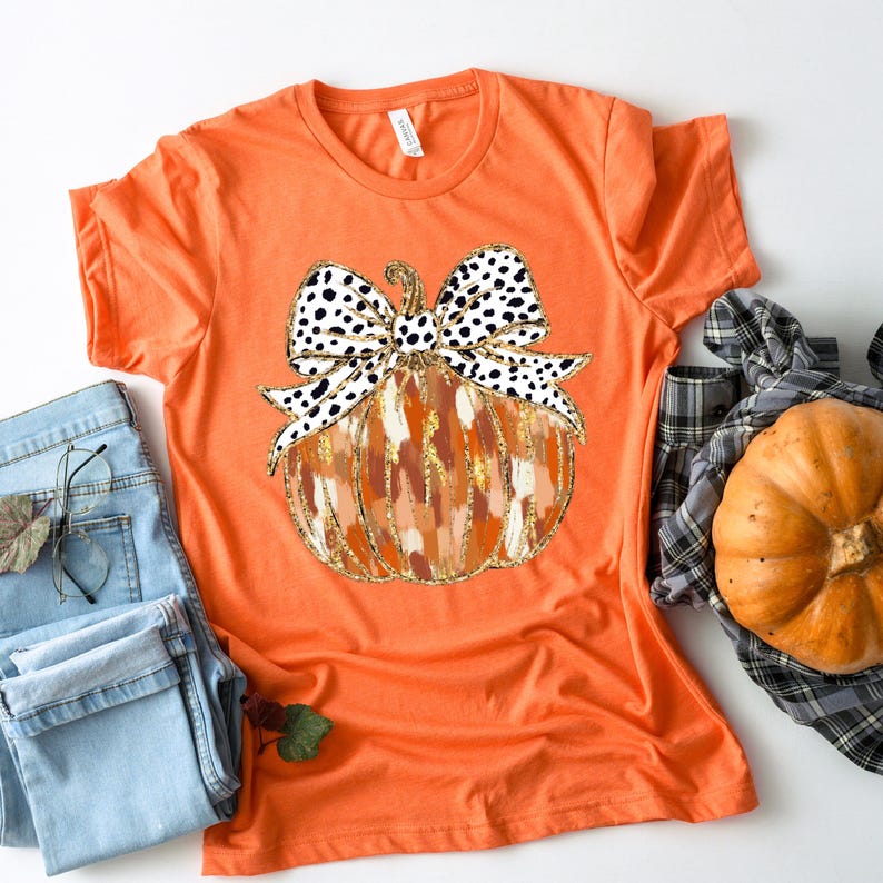 Puede incluir: Camiseta naranja con un gr&aacute;fico de una calabaza con un lazo de lunares negros y blancos. La calabaza est&aacute; pintada con pintura naranja, marr&oacute;n y dorada.
