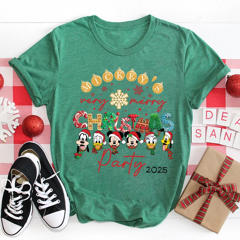 K&ouml;nnte beinhalten: Gr&uuml;nes T-Shirt mit einem Mickey Mouse Weihnachtsparty-Design. Das Design zeigt Mickey Mouse und seine Freunde in Weihnachtskost&uuml;men. Der Text auf dem Shirt lautet "Mickey's Very Merry Christmas Party 2025".