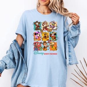 May include: Light blue t-shirt featuring a colorful graphic of Disney characters in cowboy attire. The design includes Mickey Mouse, Minnie Mouse, Chip and Dale, and Donald Duck. The text "Disney Howdy Partners" is printed below the graphic.