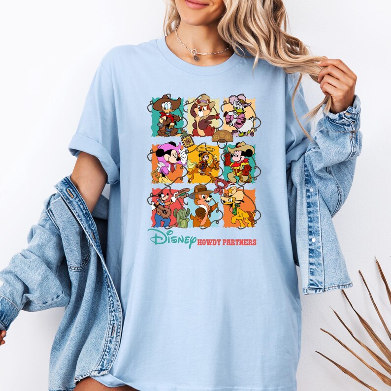 May include: Light blue t-shirt featuring a colorful graphic of Disney characters in cowboy attire. The design includes Mickey Mouse, Minnie Mouse, Chip and Dale, and Donald Duck. The text "Disney Howdy Partners" is printed below the graphic.