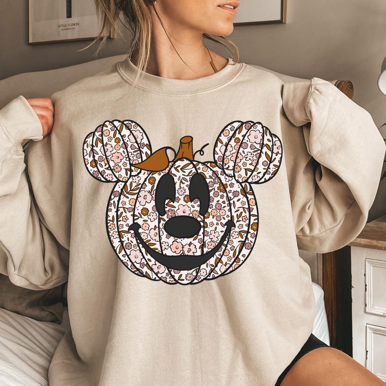 May include: A beige sweatshirt with a floral pumpkin Mickey Mouse design. The pumpkin is brown with pink and white flowers.