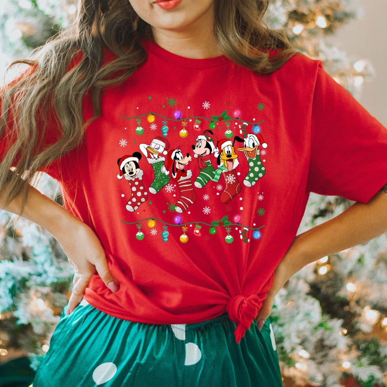 May include: Red t-shirt with a Christmas lights design featuring Mickey Mouse, Donald Duck, Goofy, Pluto, and a Christmas stocking. The characters are wearing Santa hats and are holding Christmas lights.