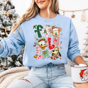 K&ouml;nnte beinhalten: Hellblaues Sweatshirt mit dem Wort "Fall", das aus weihnachtlichen Designs und Disney-Figuren besteht. Der Buchstabe "F" ist mit Stechpalmen verziert, das "A" mit Zuckerstangen, das erste "L" mit einem Weihnachtsstrumpf und das zweite "L" mit einer Glocke.