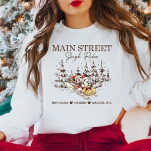 May include: White sweatshirt with a graphic featuring Mickey and Minnie Mouse on a sleigh, with pine trees in the background. Text reads "MAIN STREET Sleigh Rides" and "HOT COCOA COOKIES MAGICAL FUN".