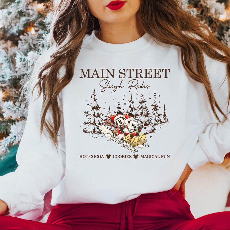 May include: White sweatshirt with a graphic featuring Mickey and Minnie Mouse on a sleigh, with pine trees in the background. Text reads "MAIN STREET Sleigh Rides" and "HOT COCOA COOKIES MAGICAL FUN".