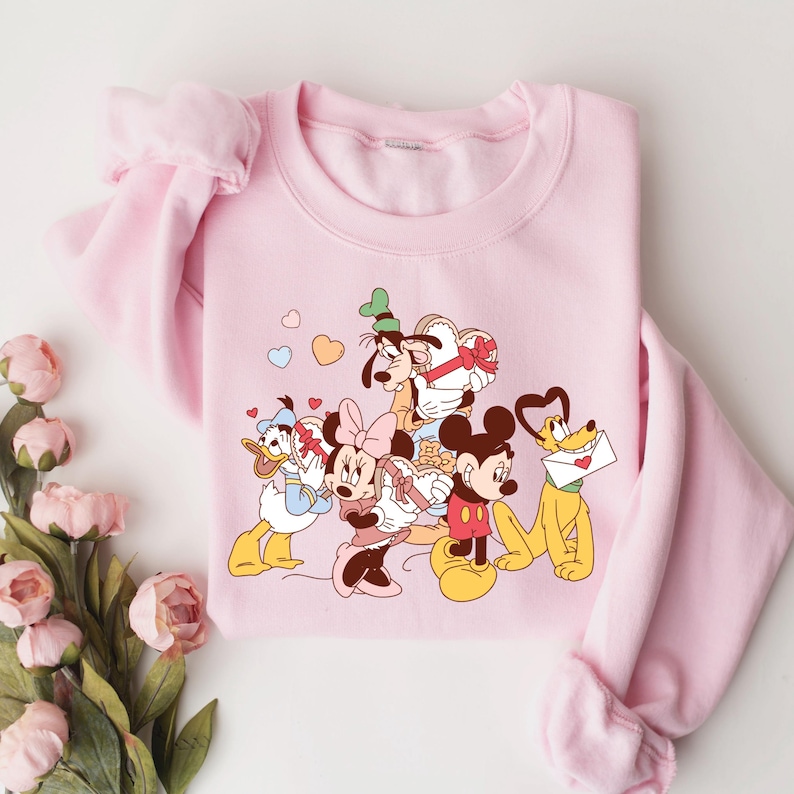 May include: Pink sweatshirt with a graphic print of Mickey Mouse, Minnie Mouse, Donald Duck, Goofy, and Pluto holding hearts and gifts. The characters are in a cartoon style and are surrounded by hearts.