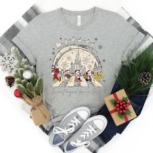 Puede incluir: Una camiseta gris con un dise&ntilde;o navide&ntilde;o que presenta a Mickey Mouse y sus amigos caminando por un paso de cebra frente al Castillo de Cenicienta. El texto "The most magical place on Earth" est&aacute; impreso debajo del dise&ntilde;o.
