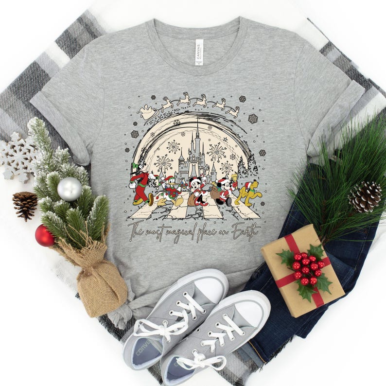Puede incluir: Una camiseta gris con un dise&ntilde;o navide&ntilde;o que presenta a Mickey Mouse y sus amigos caminando por un paso de cebra frente al Castillo de Cenicienta. El texto "The most magical place on Earth" est&aacute; impreso debajo del dise&ntilde;o.