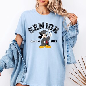 Op de afbeelding: Lichtblauw T-shirt met een zwarte afbeelding van Mickey Mouse die een afstudeerpet draagt en een diploma vasthoudt. De tekst "SENIOR" is zwart met een witte omtrek. De tekst "CLASS OF 2025" is ook zwart.