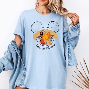 Puede incluir: Camiseta azul claro con una silueta negra de Mickey Mouse. Dentro de la silueta hay personajes de la pel&iacute;cula El Rey Le&oacute;n, incluyendo Simba, Mufasa y Pumbaa. El texto "Hakuna Matata" est&aacute; impreso debajo de la silueta.