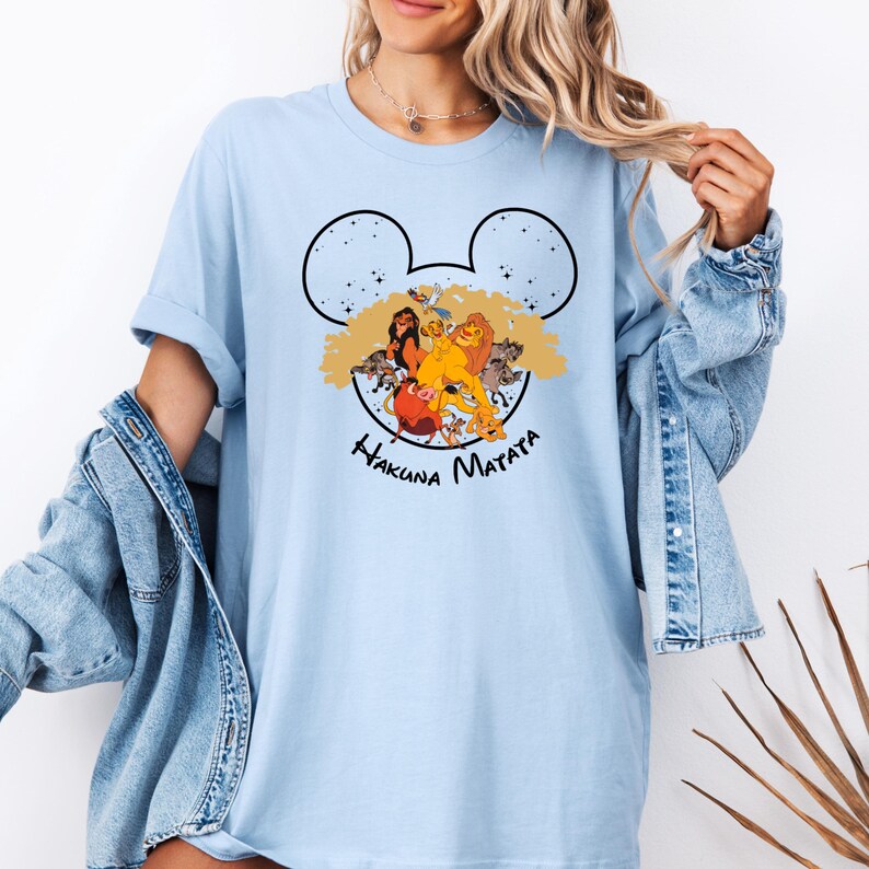 Puede incluir: Camiseta azul claro con una silueta negra de Mickey Mouse. Dentro de la silueta hay personajes de la pel&iacute;cula El Rey Le&oacute;n, incluyendo Simba, Mufasa y Pumbaa. El texto "Hakuna Matata" est&aacute; impreso debajo de la silueta.