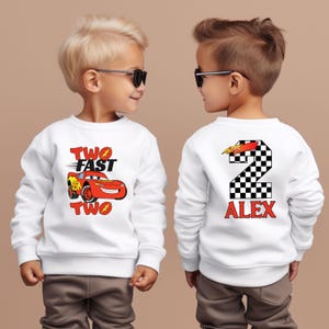 K&ouml;nnte beinhalten: Wei&szlig;es Sweatshirt mit roter Rennwagen-Grafik und dem Text "TWO FAST TWO" auf der Vorderseite. Auf der R&uuml;ckseite befindet sich eine karierte Zahl 2 mit dem Namen "ALEX" in Rot. Das Sweatshirt ist f&uuml;r ein Kind.