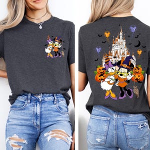 May include: Dark gray t-shirt featuring a Halloween-themed graphic of Minnie Mouse and Daisy Duck in witch costumes. The back of the shirt displays a castle, pumpkins, and bats. The shirt has a crew neck and short sleeves.