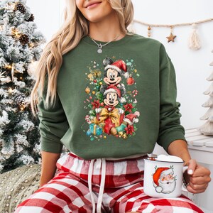 May include: Green sweatshirt featuring a festive Mickey and Minnie Mouse design wearing Santa hats, surrounded by Christmas decorations and gifts. The image also shows a Santa mug and red and white plaid pajama pants.