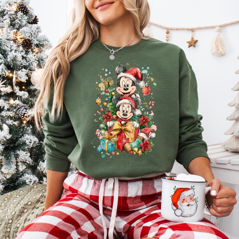 May include: Green sweatshirt featuring a festive Mickey and Minnie Mouse design wearing Santa hats, surrounded by Christmas decorations and gifts. The image also shows a Santa mug and red and white plaid pajama pants.