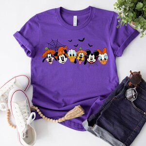 May include: Purple t-shirt with a graphic of Mickey Mouse, Minnie Mouse, Donald Duck, Goofy, and Pluto dressed in Halloween costumes. The characters are lined up in a row with a spider web and bats in the background.