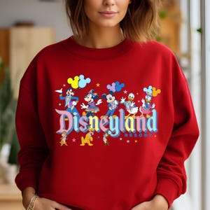 May include: Red sweatshirt with a colourful Disneyland graphic featuring Mickey Mouse, Minnie Mouse, Donald Duck, Goofy, and Pluto. The graphic includes the text "Disneyland Resort".