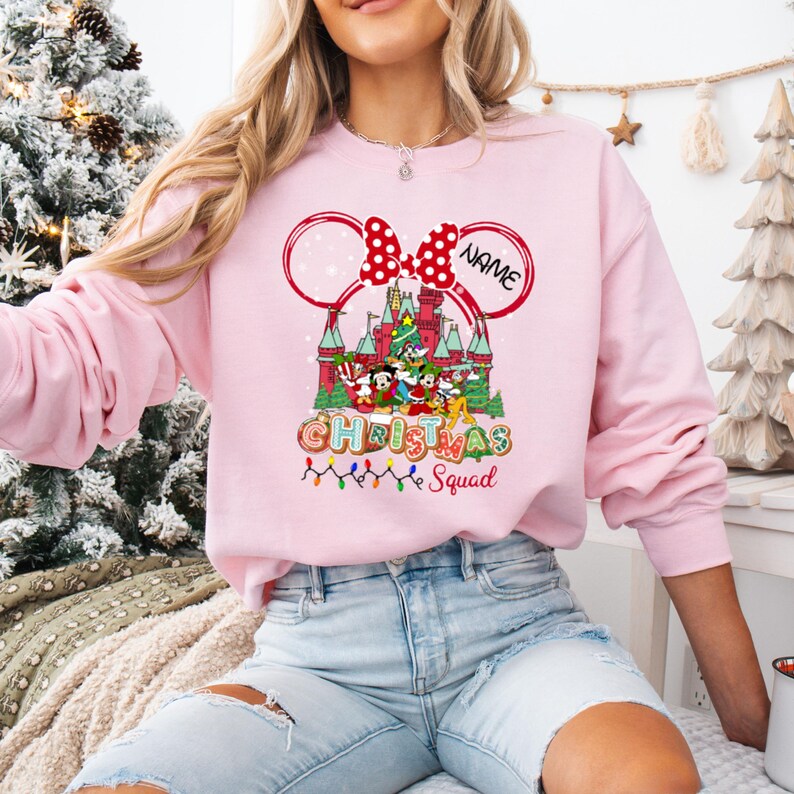 Puede incluir: Sudadera rosa claro con un dise&ntilde;o navide&ntilde;o. El gr&aacute;fico incluye un castillo, personajes de Disney y las palabras "Christmas Squad". Un lazo rojo y orejas de Mickey Mouse enmarcan el dise&ntilde;o. La palabra "NAME" est&aacute; escrita en la oreja derecha.