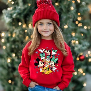 K&ouml;nnte beinhalten: Ein junger Mensch tr&auml;gt ein rotes Sweatshirt mit Mickey Mouse, Minnie Mouse, Donald Duck, Daisy Duck, Goofy und Pluto. Das Sweatshirt hat ein festliches Design mit Lichterketten und Schneeflocken. Die Person tr&auml;gt auch eine rote Strickm&uuml;tze.