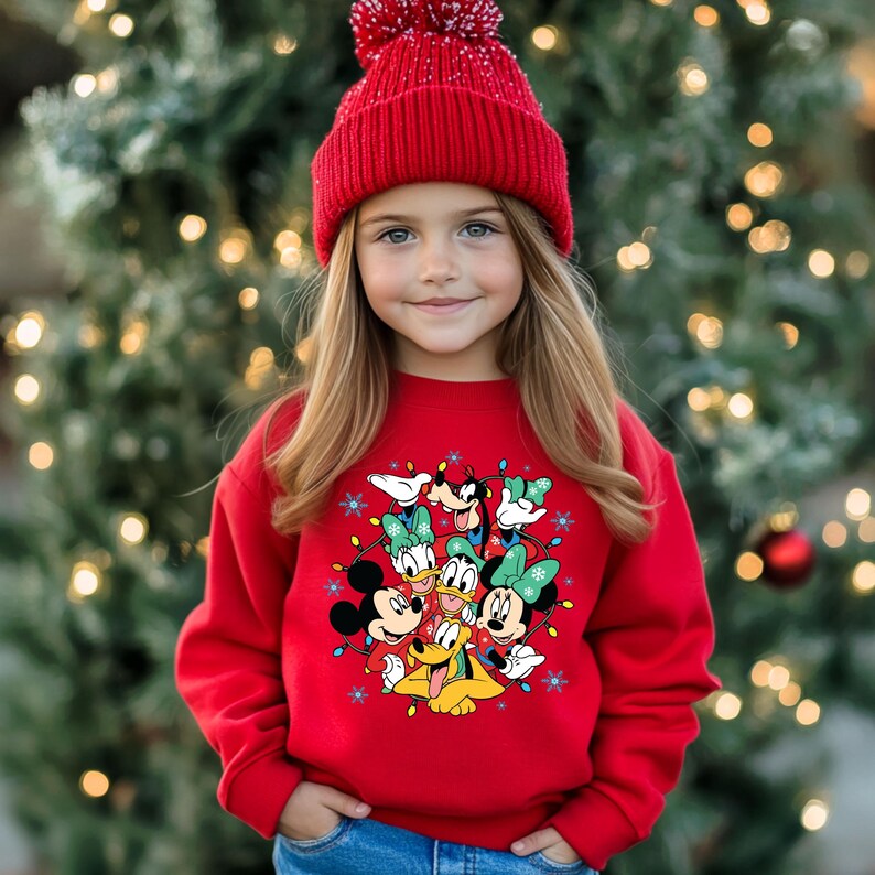 K&ouml;nnte beinhalten: Ein junger Mensch tr&auml;gt ein rotes Sweatshirt mit Mickey Mouse, Minnie Mouse, Donald Duck, Daisy Duck, Goofy und Pluto. Das Sweatshirt hat ein festliches Design mit Lichterketten und Schneeflocken. Die Person tr&auml;gt auch eine rote Strickm&uuml;tze.