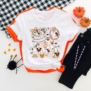 May include: White t-shirt with a Halloween-themed graphic featuring cartoon ghosts, pumpkins, and the text "HAPPY HALLOWEEN." The design includes Mickey and Minnie Mouse characters in ghost costumes. The shirt is paired with black jeans and orange accents.