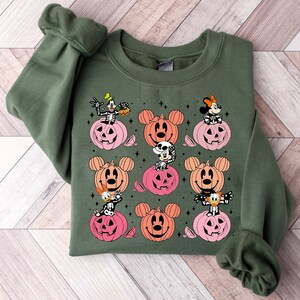 May include: Green sweatshirt with a Halloween-themed design featuring Mickey Mouse, Minnie Mouse, Goofy, and Donald Duck characters in pumpkin costumes. The characters are depicted in a variety of poses, with some holding candy corn and others smiling.