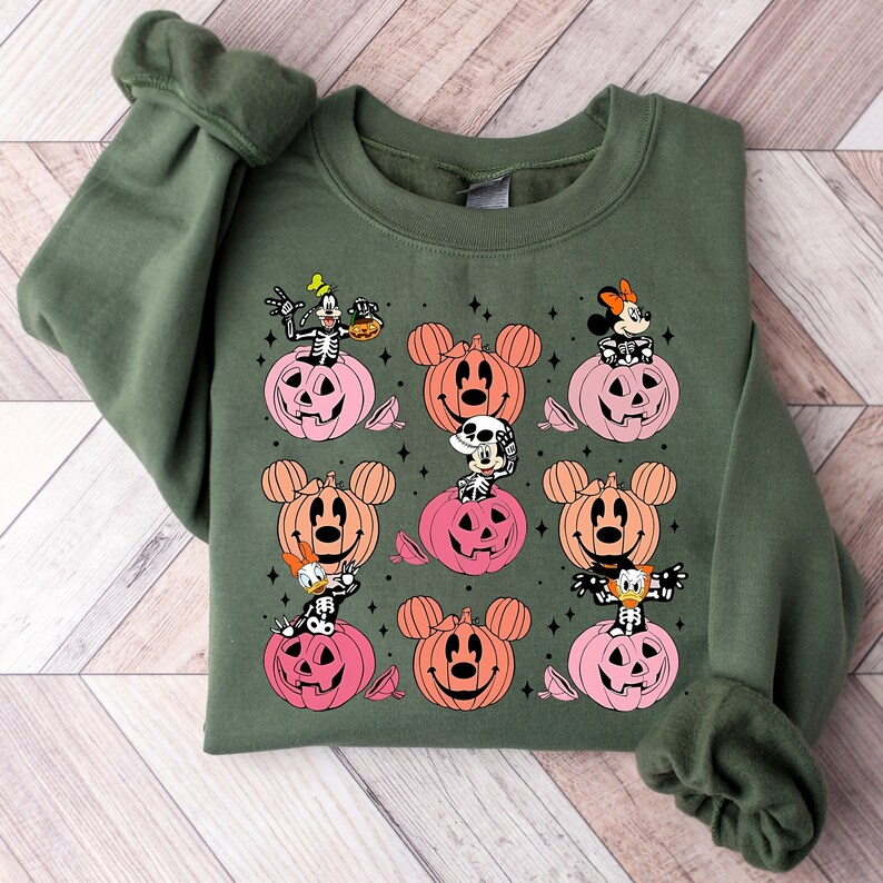 May include: Green sweatshirt with a Halloween-themed design featuring Mickey Mouse, Minnie Mouse, Goofy, and Donald Duck characters in pumpkin costumes. The characters are depicted in a variety of poses, with some holding candy corn and others smiling.
