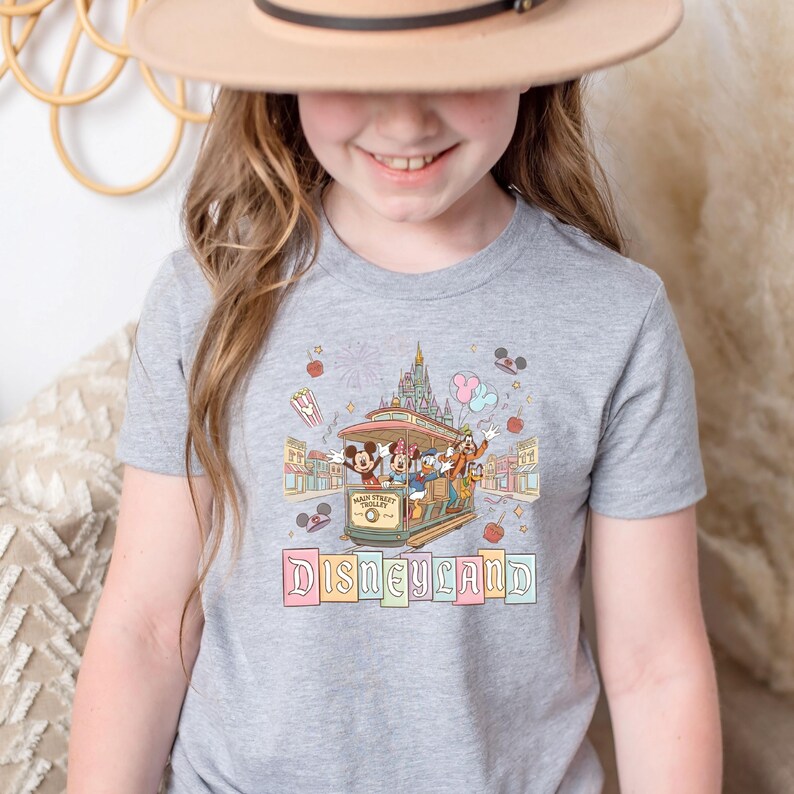 May include: A gray t-shirt featuring a colorful Disneyland graphic. The design includes a trolley with Mickey Mouse, Donald Duck, and Goofy, a castle, and the word "Disneyland" in pastel letters. The shirt is likely for children.