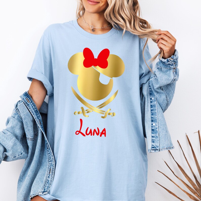 May include: Light blue t-shirt with a gold Mickey Mouse head silhouette with a red bow and an eye patch. Two gold crossed swords are below the head. The name "Luna" is written in red below the swords.