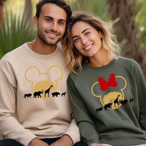 May include: A couple wearing matching sweatshirts with a Mickey Mouse silhouette design featuring a safari theme. The sweatshirts are beige and olive green with black animal silhouettes and a red bow.