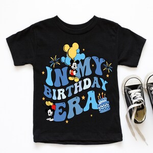 May include: Black t-shirt with a blue and yellow graphic of Mickey Mouse celebrating his birthday. The text on the shirt reads "In My Birthday Era".