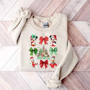 May include: A beige sweatshirt with a Christmas-themed design featuring Mickey Mouse, Minnie Mouse, Donald Duck, and a depiction of the Disney castle. The design includes red and green bows, snowflakes, and a green and white plaid pattern.