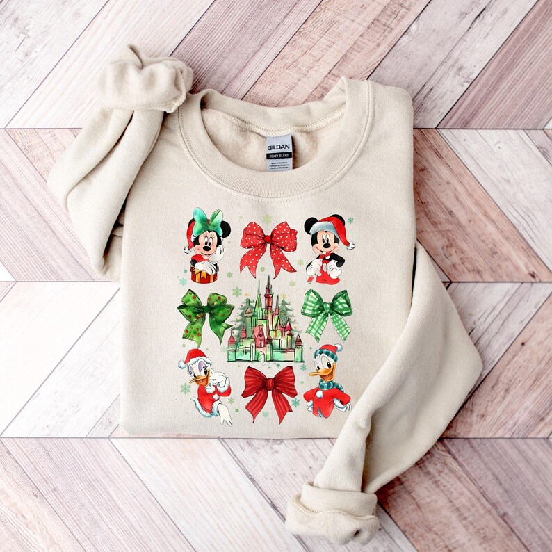 May include: A beige sweatshirt with a Christmas-themed design featuring Mickey Mouse, Minnie Mouse, Donald Duck, and a depiction of the Disney castle. The design includes red and green bows, snowflakes, and a green and white plaid pattern.