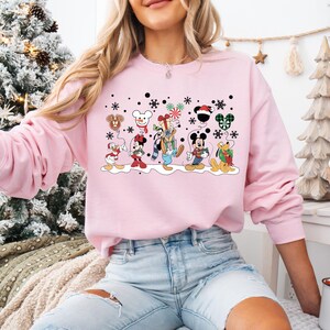 May include: Pink Christmas sweatshirt featuring a group of Disney characters including Mickey Mouse, Minnie Mouse, Donald Duck, Goofy, and Pluto. The characters are wearing winter clothing and holding gifts and balloons.