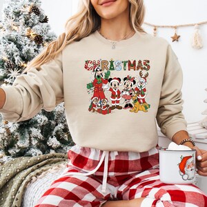 May include: Beige Christmas sweatshirt featuring a colorful graphic of Disney characters, including Mickey and Minnie Mouse, Goofy, and Pluto. The word "CHRISTMAS" is written across the top in festive lettering. The sweatshirt is paired with red and white plaid pajama pants.