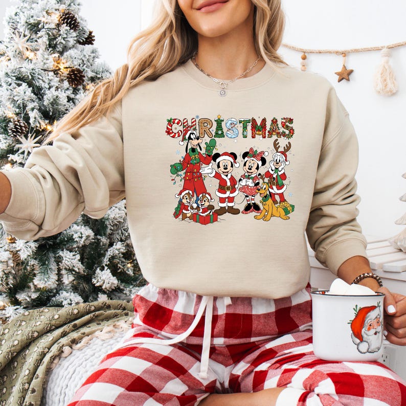 May include: Beige Christmas sweatshirt featuring a colorful graphic of Disney characters, including Mickey and Minnie Mouse, Goofy, and Pluto. The word "CHRISTMAS" is written across the top in festive lettering. The sweatshirt is paired with red and white plaid pajama pants.