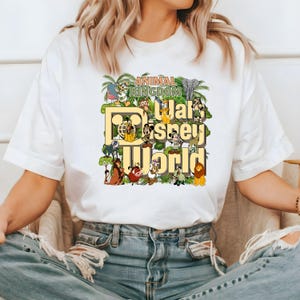 Pu&ograve; includere: Maglietta bianca con un design grafico colorato che presenta il testo "Animal Kingdom Walt Disney World" e vari personaggi e animali Disney.