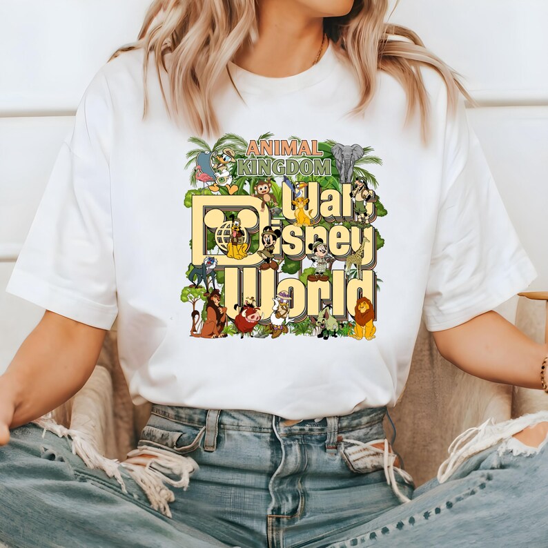 Pu&ograve; includere: Maglietta bianca con un design grafico colorato che presenta il testo "Animal Kingdom Walt Disney World" e vari personaggi e animali Disney.