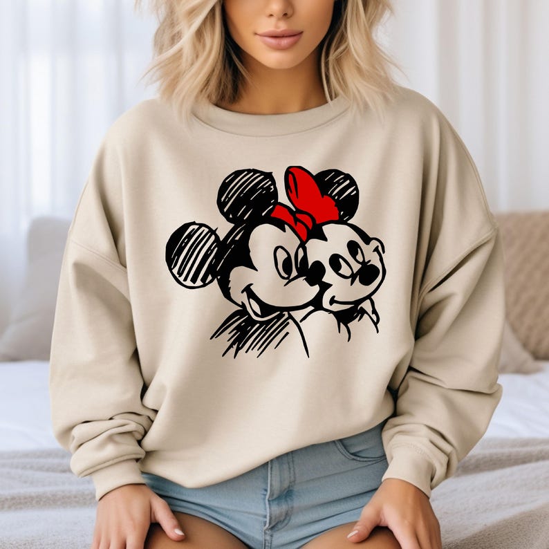 May include: Beige sweatshirt featuring a black and red graphic of Mickey and Minnie Mouse. The cartoon characters are drawn in a sketch style, with Mickey's ears and Minnie's bow in red. The sweatshirt is a neutral color.