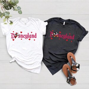 May include: Two t-shirts with a pink Disneyland logo and Mickey Mouse and Minnie Mouse graphics. The white t-shirt has red hearts around the logo. The black t-shirt has red hearts above the logo.