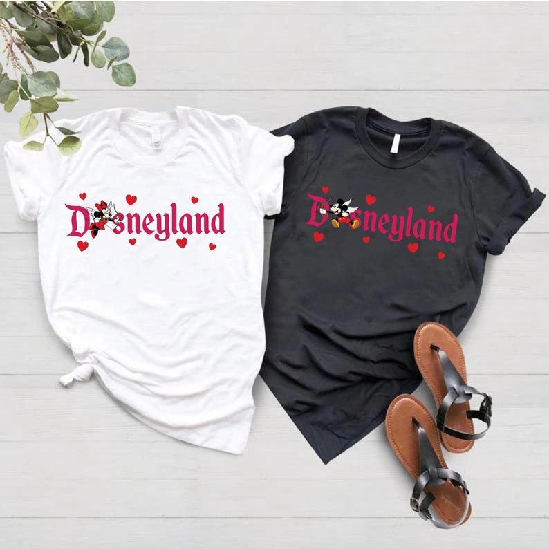 May include: Two t-shirts with a pink Disneyland logo and Mickey Mouse and Minnie Mouse graphics. The white t-shirt has red hearts around the logo. The black t-shirt has red hearts above the logo.