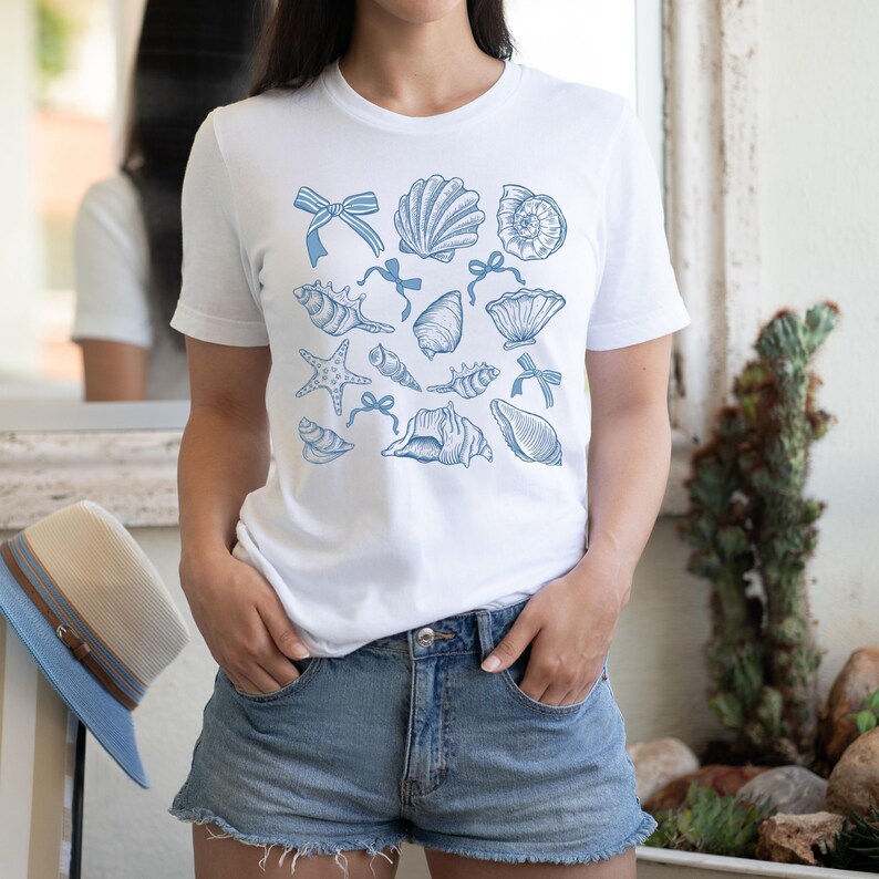 Pu&ograve; includere: Maglietta bianca con un motivo blu di conchiglie e stelle marine. Il design presenta varie conchiglie, stelle marine e fiocchi.
